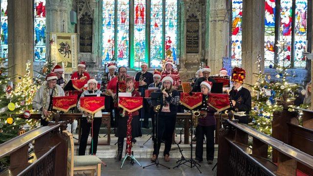 Saxophonics band, standing playing a selection of saxophones, whearing santa hats and tinsel in front of the church altar